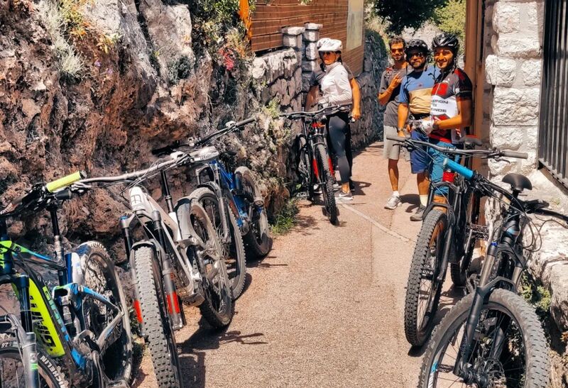 Des VTT électriques alignés le long d'un mur de pierre dans une ruelle de Peille, avec un groupe de cyclistes prêts à partir