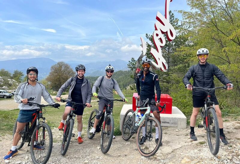 Un groupe de cinq cyclistes et leurs vélos devant une sculpture rouge et blanche, sommets au loin